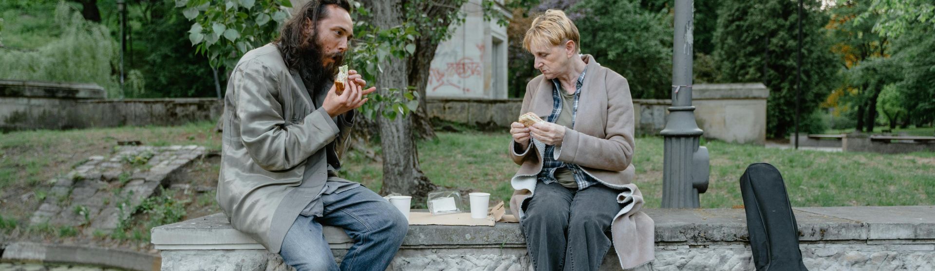 Two individuals sharing a meal outdoors on a stone bench, highlighting companionship and resilience.